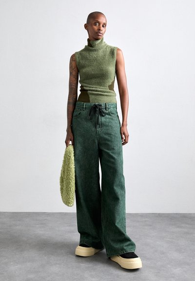 Πράσινο αμάνικο πουλόβερ με ψηλό γιακά συνδυασμένο με φαρδιά πράσινα τζιν παντελόνια. Διαθέτει μαύρη ελαστική ζώνη και κρεμ πλατφόρμες. Κρατάει μια υφασμάτινη πράσινη τσάντα.