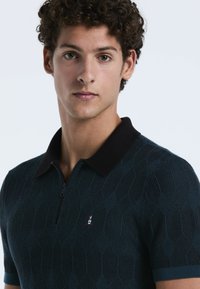 Jonge man met krullend donker haar draagt een donkergroen gestructureerd polo-shirt met een zwarte kraag en een klein geborduurd logo op de borst.