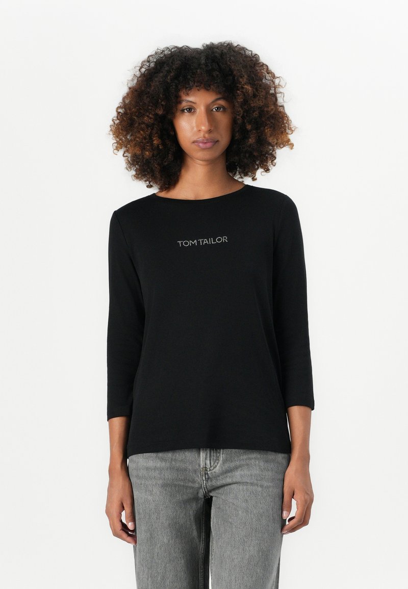Black three-quarter sleeve shirt with round neckline and embroidered "TOM TAILOR" logo on the chest, paired with gray straight-leg jeans.