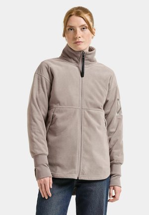 Giovane donna con capelli rossi che indossa una giacca in pile beige con collo alto e cerniera completa, abbinata a jeans blu, in piedi su sfondo bianco.