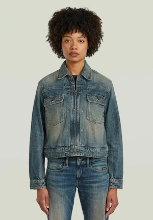 Vrouw met krullend haar draagt een verwassen blauw spijkerjack half dicht en bijpassende high-waisted jeans, staand tegen een effen achtergrond.