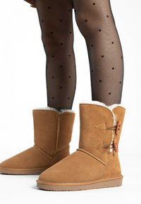 Tan suède enkellaarsjes hebben een zachte witte bontvoering, lusriemen met houten knopen en een platte rubberen zool met grip.
