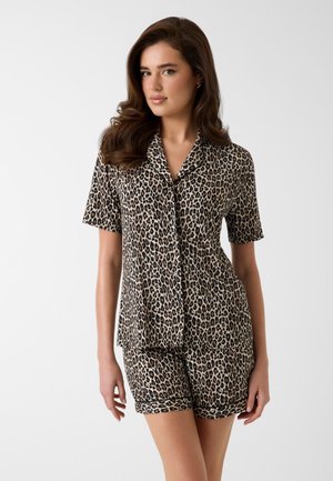 Joven mujer con camisa de manga corta con estampado de leopardo y pantalones cortos a juego sobre un fondo claro y liso.