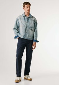Jeune homme debout, une main dans la poche, portant une veste en denim bleu clair, un pantalon bleu marine et des baskets blanches à semelles marron.