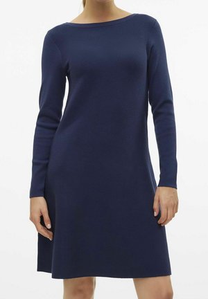 Kvinde iført en navyblå, strikket kjole med lange ærmer og knælængde med bådformet udskæring, stående mod en enkel hvid baggrund.