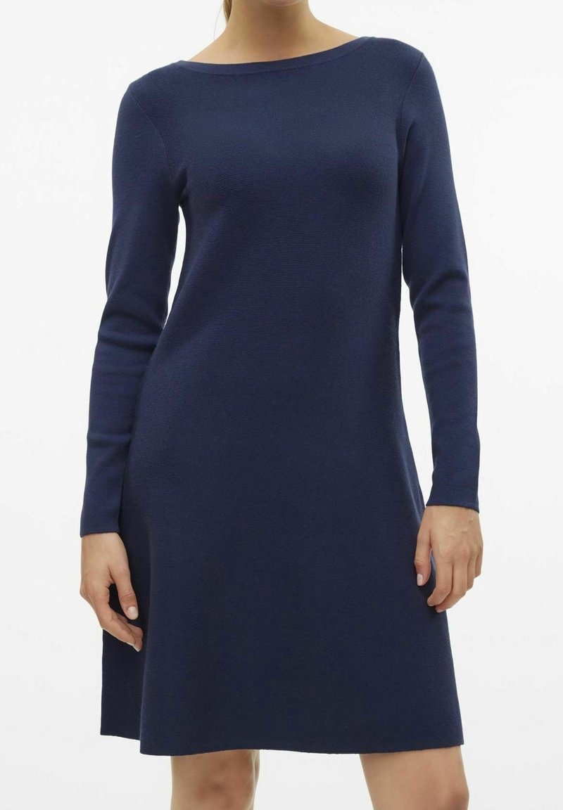 Femme portant une robe en maille bleu marine à manches longues, longueur genou, avec un col bateau, debout devant un fond blanc uni.