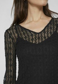 Melna mežģīņu blūze ar garām piedurknēm, kurai ir V veida dekoltē un struktūras raksti ar kombināciju no necaurspīdīgas un caurspīdīgas daļām.