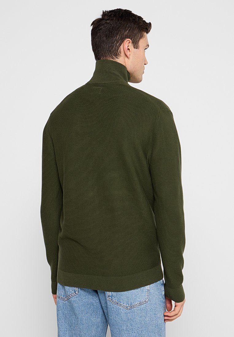 Mann mit kurzem dunklem Haar, der einen olivgrünen Rollkragenpullover und hellblaue Jeans trägt, mit dem Rücken zu einem schlichten hellen Hintergrund gewandt.