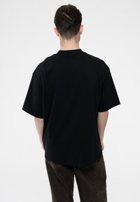 Black cotton t-shirt with a relaxed fit, short sleeves, and a round neckline. Seam detailing at the back, worn with dark brown pants.