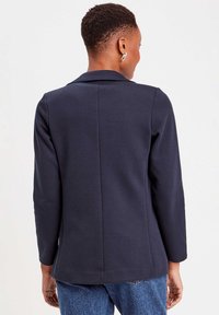 Marineblauwe blazer in een gestructureerd ontwerp, met een opstaande revers en lange mouwen. Glad materiaal, enkele rugnaad, gedragen over lichtblauwe jeans.