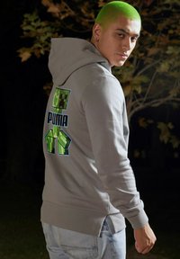 Lys grå hættejakke lavet af bomuld med en afslappet pasform, prydet med et grønt pixeliseret grafik af en karakter og "PUMA"-tekst på ryggen.