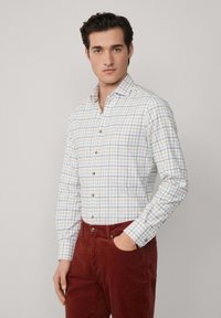 Camicia bianca con un motivo a griglia blu e arancione. Presenta un colletto, maniche lunghe e bottoni marroni. Abbinata a pantaloni bordeaux.