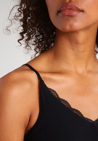 Musta toppi ohutolkaimilla, jossa on hieno pitsireunus kaula-aukossa. Sujuva pinta ja V-muotoinen kaula-aukko. Ihon sävyt näkyvissä.