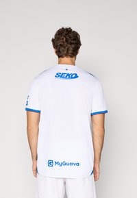 Balts sporta krekls ar zilām akcentiem un sarkanu apdari. Tālajā augšējā daļā redzams zils "SEKO" logo, bet tuvu apakšai atrodams "MyGuava" logo.