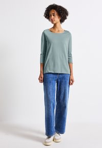 Top verde chiaro a maniche lunghe con scollatura rotonda, abbinato a jeans a gamba larga blu e sneakers beige. Il tessuto sembra morbido e casual.