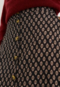 Jupe noire avec un motif floral doré, en tissu texturé, et boutons en laiton sur le côté, associée à un pull rouge.