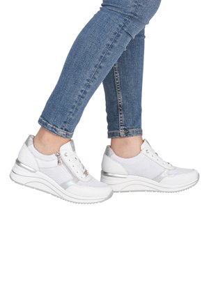 Jambes portant un jean skinny bleu et des baskets blanches de sport avec des fermetures éclair latérales et des semelles épaisses, présentées sur un fond blanc.