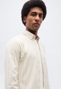 Homme avec un afro court portant une chemise à boutons à rayures verticales beige et blanche par-dessus un t-shirt blanc, sur un fond clair uni.
