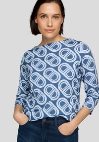 Blaues Langarmhemd mit einem sich wiederholenden weißen und hellblauen kreisförmigen Logomuster, rundem Ausschnitt und weicher Textur, kombiniert mit dunklen Jeans.