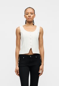 Νεαρό άτομο με πλεξούδες, ντυμένο με άσπρο αμάνικο τοπ με κουμπιά και μαύρο παντελόνι, στέκεται μπροστά σε απλό λευκό φόντο.