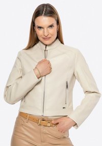 Giacca cropped in pelle crema con colletto alto, chiusura frontale a zip e tasche laterali. Texture liscia con dettagli di cucitura e pantaloni in tonalità neutra.