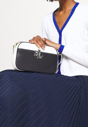 Vrouw in een witte cardigan met blauwe bies houdt een zwart-witte leren crossbodytas met "RLL"-logo vast, draagt een marineblauwe geplooide rok.