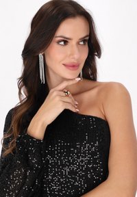 Schwarzes, glitzerndes Off-Shoulder-Oberteil mit puffigen Ärmeln. Das Model trägt silberne, hängende Ohrringe und einen runden Goldring.