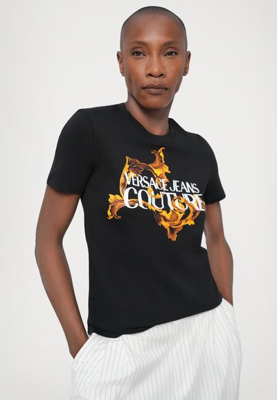 T-shirt noir à manches courtes avec un motif floral orange et or audacieux et le texte "Versace Jeans Couture" en blanc. Matière en coton, design ajusté.