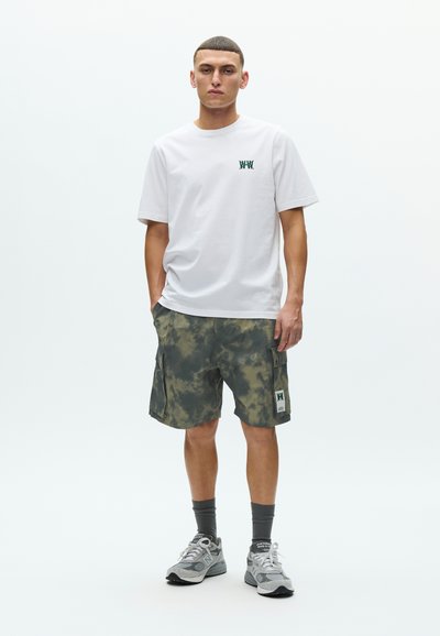 Λευκό βαμβακερό t-shirt με πράσινο λογότυπο, συνδυασμένο με πράσινα cargo σορτς tie-dye που διαθέτουν δύο πλαϊνές τσέπες και σημείο με λογότυπο. Γκρι παπούτσια για τρέξιμο.