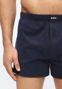 Homme portant un boxer bleu marine à taille élastique avec braguette à boutons et étiquette "BOSS" sur la ceinture.