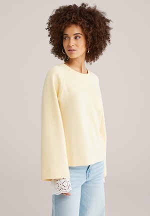 Göndör hajú nő halvány sárga, hosszú ujjú felsőt visel fehér csipkés mandzsettával és világoskék farmert, oldalra néz.