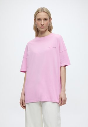 Kobieta o blond włosach do ramion, ubrana w luźny jasnoróżowy T-shirt z napisem "keep on moving" oraz białe spodnie o szerokich nogawkach, stojąca.