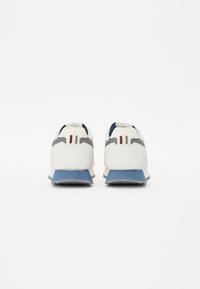 Vita och grå sneakers med blå sula, texturerad mocka och slät läderfinish, med två röda ränder på hälen.