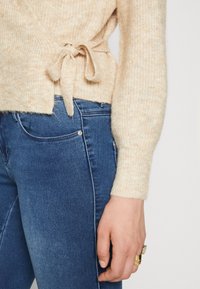Jasno-beżowy dzianinowy kardigan z detalem w postaci paska w talii, w zestawieniu z ciemnoniebieskimi dopasowanymi dżinsami. Materiał wydaje się być miękki i teksturowany.