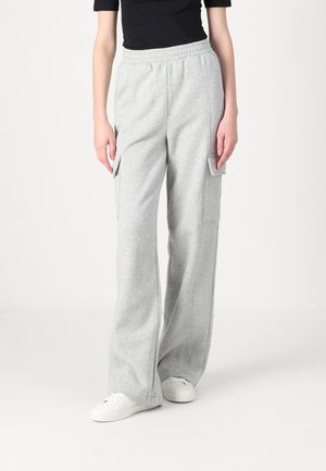 Person iført lysegrå vidtbenede joggingbukser med cargolommer, sort top og hvide sneakers, stående foran en ensfarvet baggrund.