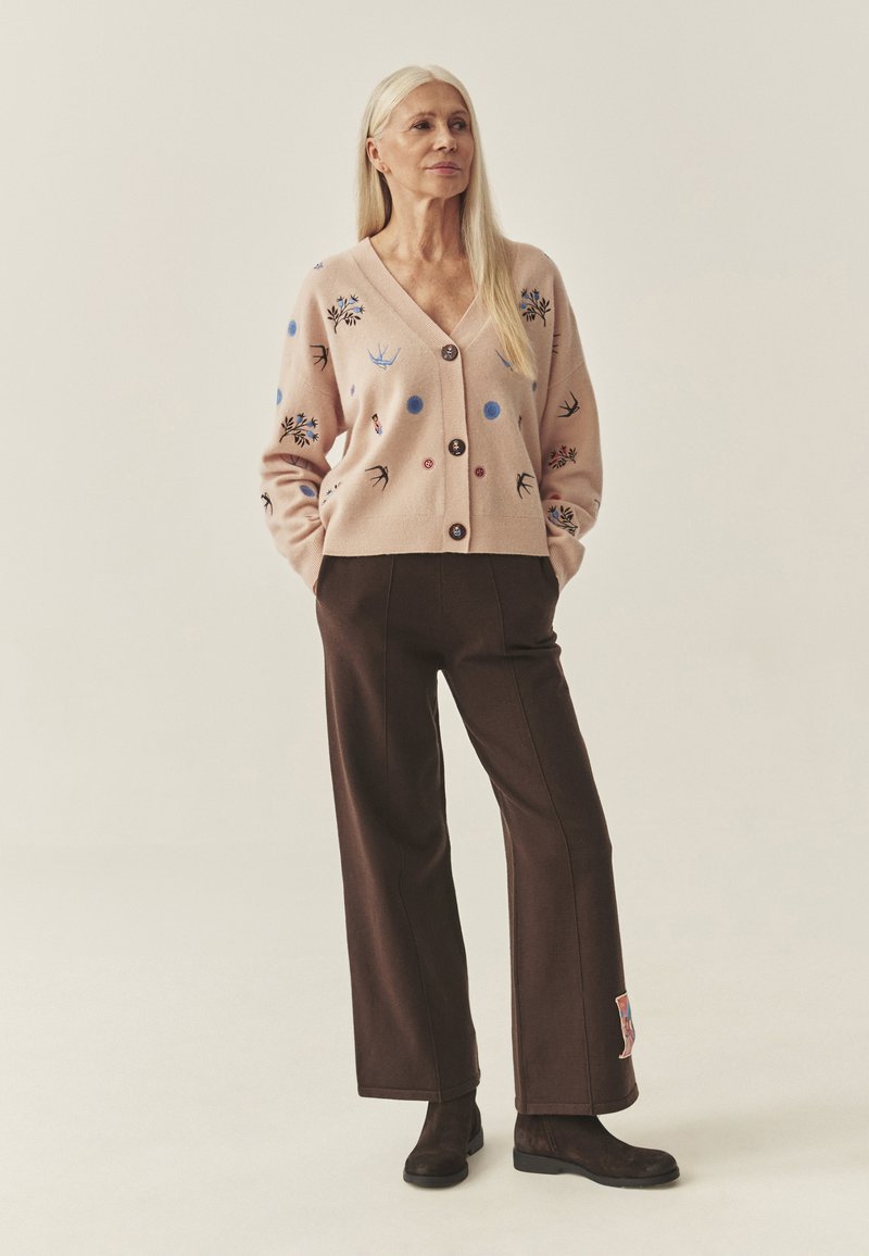 Lichtroze cardigan met bloemenborduurwerk, knopen in het midden, gecombineerd met donkerbruine wijde broek en enkellaarsjes. Soepele textuur.