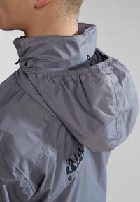 Graue wasserdichte Jacke mit Kapuze und elastischem Saum, Druckknopfverschlüssen und einem Logo auf der Rückseite, die eine glatte, glänzende Textur zeigt.