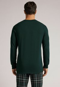 Długi rękaw, ciemnozielona koszula z okrągłym dekoltem, gładka tekstura, dopasowane rękawy, w parze z spodniami w kratę w ciemnych i jasnych kolorach.