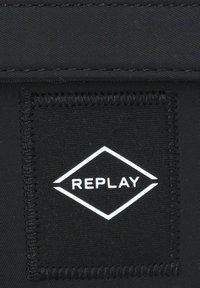 Schwarz-textiletikett mit einer Diamantform und "REPLAY" in weißer Schrift. Auffällige Naht um die Ränder und strukturierte Oberfläche.
