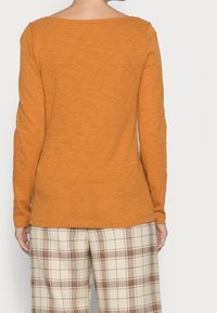 Maglia a maniche lunghe in arancio bruciato, con scollatura rotonda e morbida al tatto, abbinata a pantaloni a quadri beige con accenti arancioni.