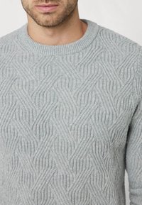 Pull en tricot gris clair avec un motif texturé en vagues, un col rond et des poignets côtelés. Tissu doux avec une apparence chaleureuse.
