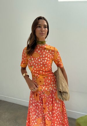 Kvinne med langt brunt hår iført en oransje kjole med hvite prikker og store gule blomster, accessorert med store ringøredobber og en beige veske.