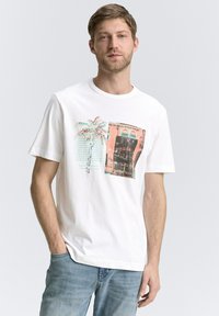 Hvid bomulds t-shirt med palmetræer og arkitektoniske prints i kontrasterende farver. Rund hals og korte ærmer, afslappet pasform.