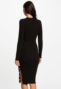 Abito nero a costine con maniche lunghe, spacco laterale e rifiniture con bottoni vicino all'orlo. Fit aderente, raggiunge la lunghezza a metà polpaccio.