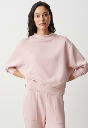 Donna con lunghi capelli scuri che indossa una felpa oversize rosa chiaro e pantaloni abbinati, in piedi con le mani sui fianchi davanti a uno sfondo semplice.