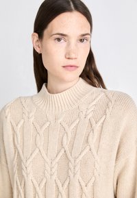 Beige knitted sweater with a high ribbed collar, featuring a cable knit pattern throughout. Soft texture and casual fit.