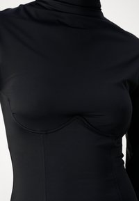 Svart långärmad topp med en mock turtleneck. Har en figursydd design och sömdetaljer över bröstområdet, tillverkad av mjukt, elastiskt tyg.