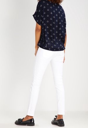 Mujer vestida con top azul marino de manga corta con anclas blancas, pantalones blancos ceñidos y mocasines negros, de pie de espaldas sobre un fondo liso.