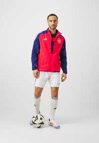 Chaqueta deportiva roja y marina con cremallera frontal, logo en el pecho; combinada con pantalones cortos blancos, calcetines y botas de fútbol con acentos azules, sosteniendo un balón.
