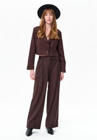 Vrouw met donkerbruine cropped blazer met krijtstreep, wijde broekspijpen, zwarte spitse laarzen en zwarte hoed met brede rand, staand tegen een witte achtergrond.